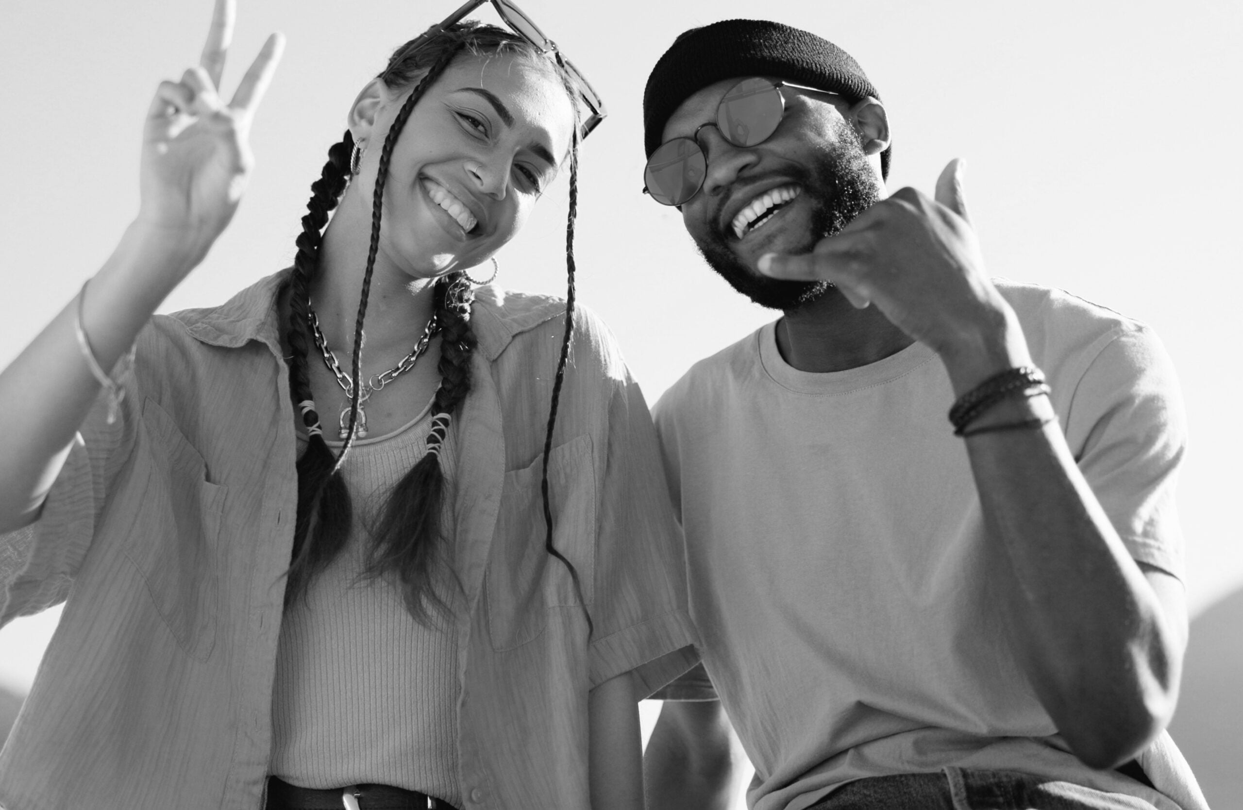 Black and white image of a girl with braids and a guy wearing a hat and sunglasses smiling 